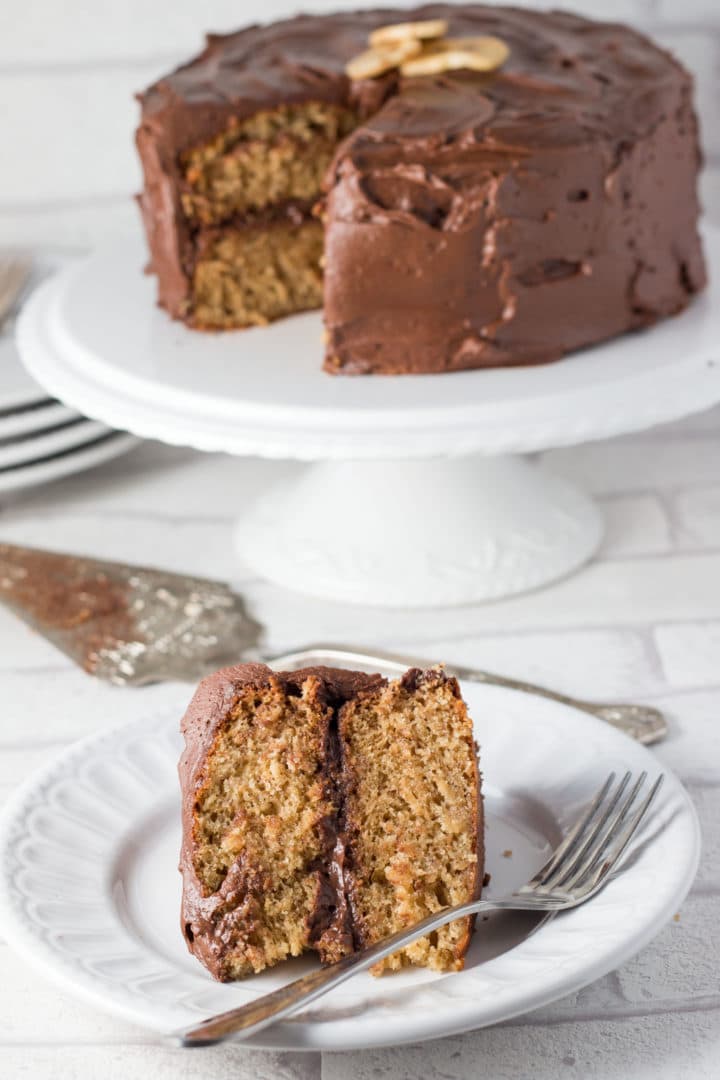 A slice of Banana Spice Cake with Chocolate Fudge Frosting on a plate with a spoon, with a cake slice and the remaining cake on a cake stand in the background