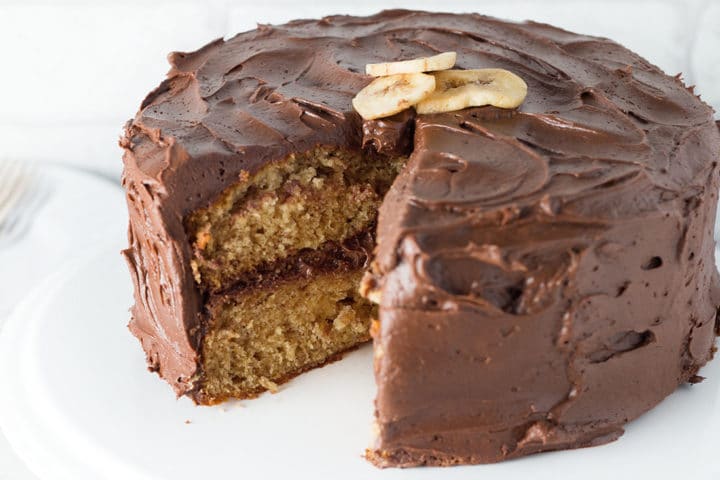 Banana Spice Cake with Chocolate Fudge Frosting with a piece taken out of it on a white background