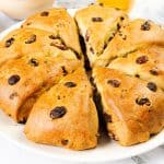 A round loaf of golden-brown Raisin Scones, sliced into eight wedges, is displayed on a white plate. Bowls of raisins and a cup are visible in the background.