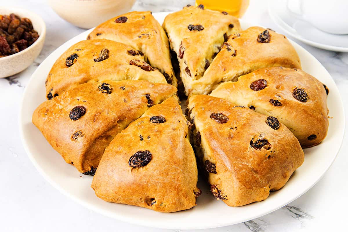 A round loaf of golden-brown Raisin Scones, sliced into eight wedges, is displayed on a white plate. Bowls of raisins and a cup are visible in the background.