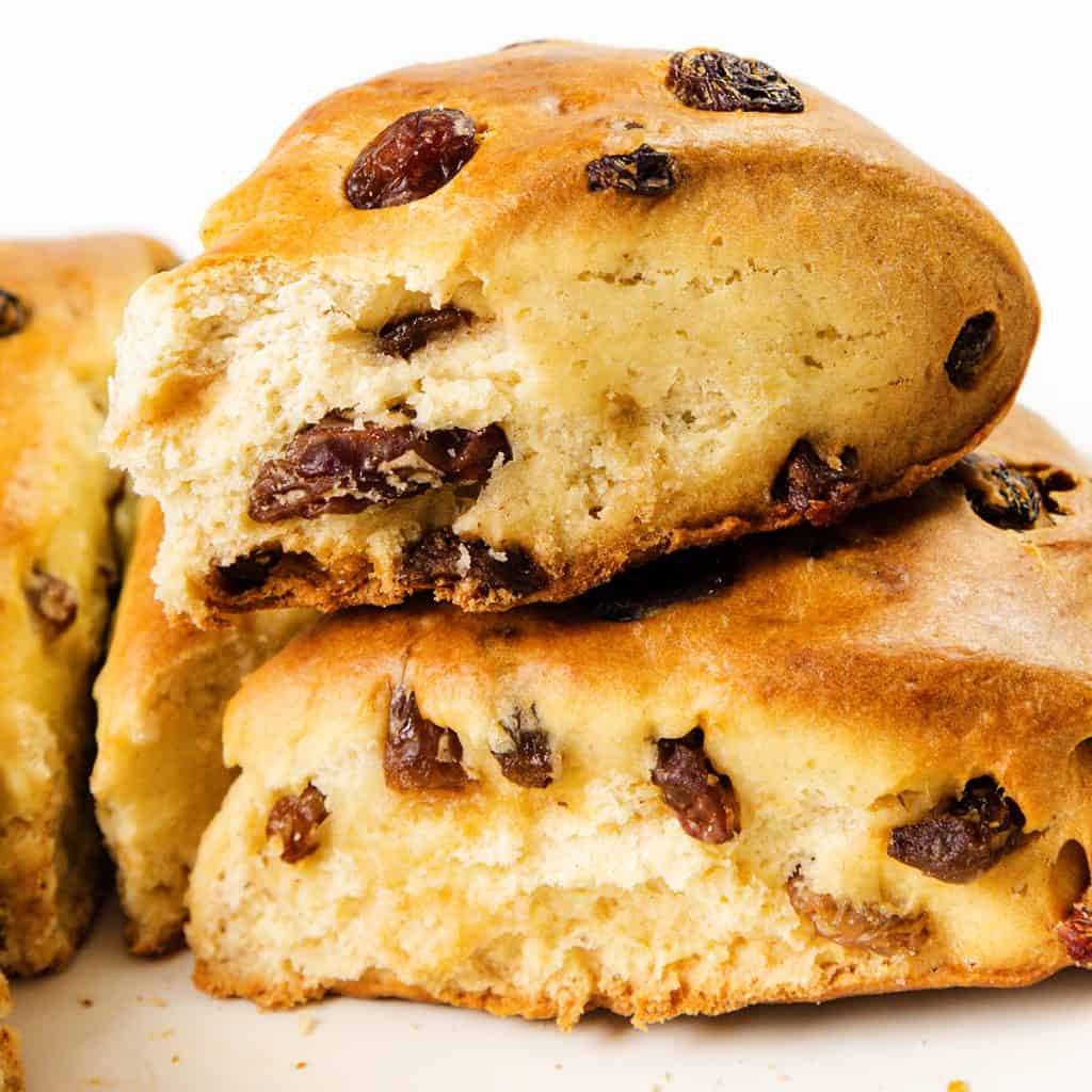 Two thick, golden-brown raisin scones are stacked on a white surface. The top scone has a bite taken out, revealing a soft, fluffy interior packed with juicy raisins.