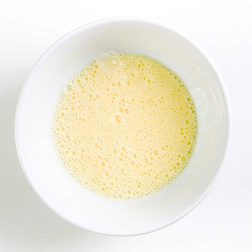 A white bowl filled with a light yellow, frothy liquid, likely beaten eggs or an egg mixture for raisin scones, viewed from above on a white background.