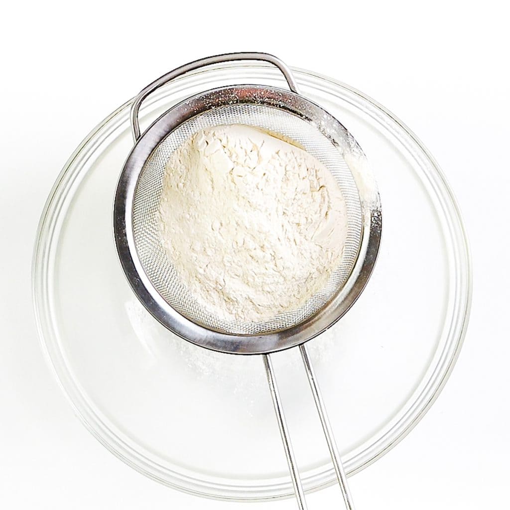 A metal sifter containing flour rests over a clear glass bowl, with some flour already sifted into the bowl below—perfectly prepped for baking delicious Raisin Scones, all set against a white background.