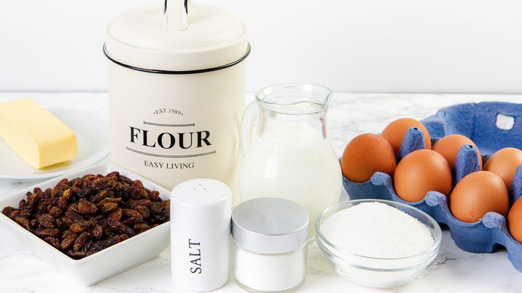 Baking ingredients on a counter: a flour canister, a glass pitcher of milk, a plate of butter, a salt shaker, sugar, raisins, and a blue egg carton with brown eggs—everything you need to make delicious raisin scones.