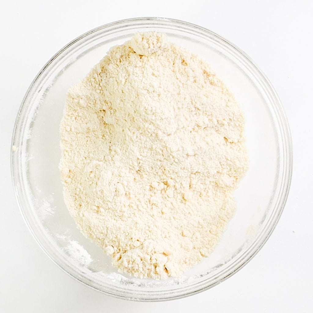 A clear glass bowl filled with an off-white powder, likely flour for Raisin Scones, sits on a white surface.