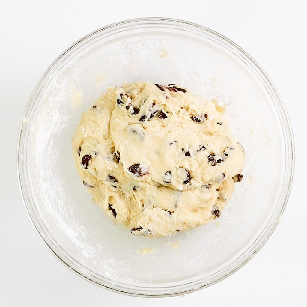 A glass bowl containing dough for raisin scones, mixed with plump raisins, resting on a white surface.