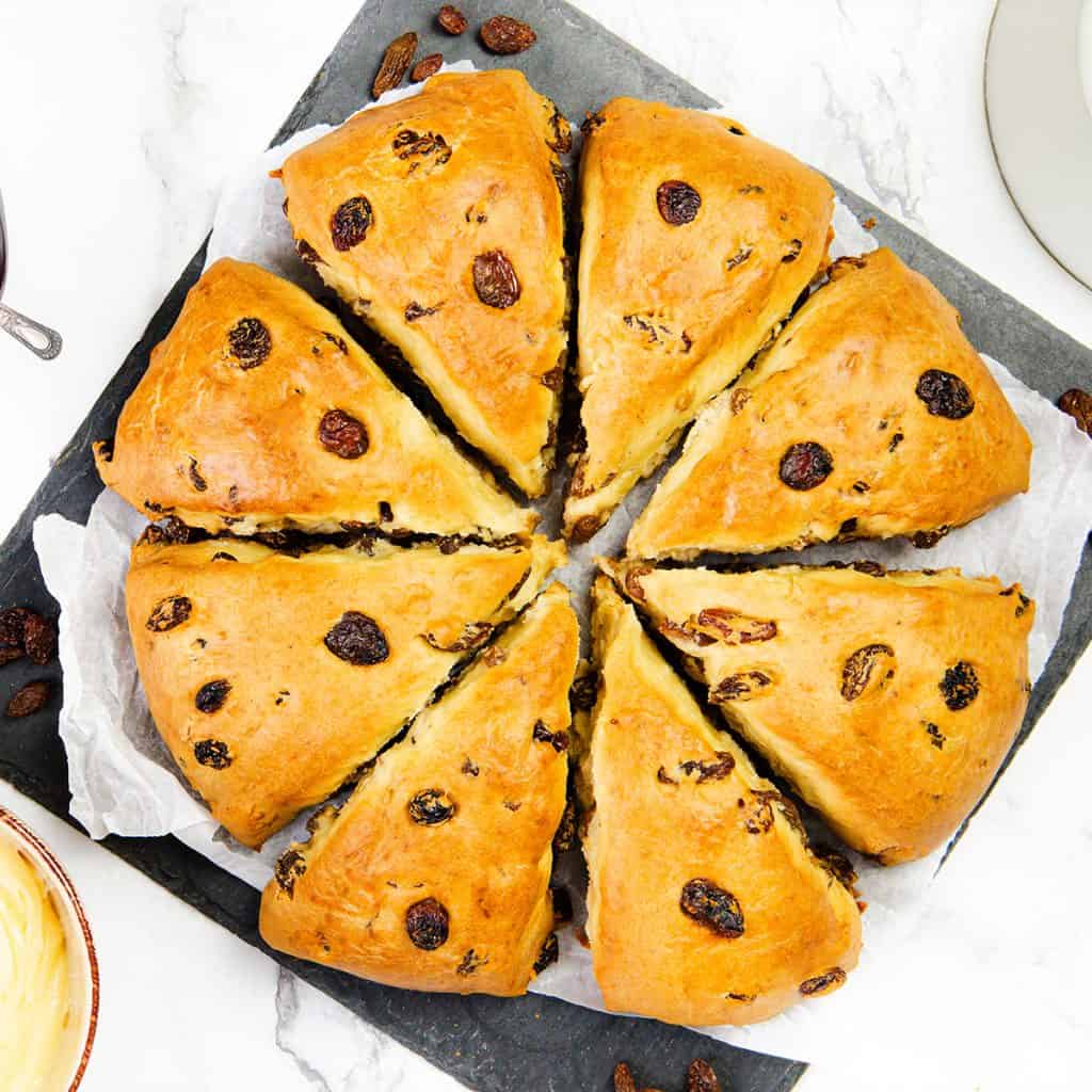 A round, golden-brown Raisin Scones loaf is cut into eight triangular slices, arranged in a circle on baking parchment atop a dark slate board. A dish of butter and scattered raisins rest nearby on a white marble surface.