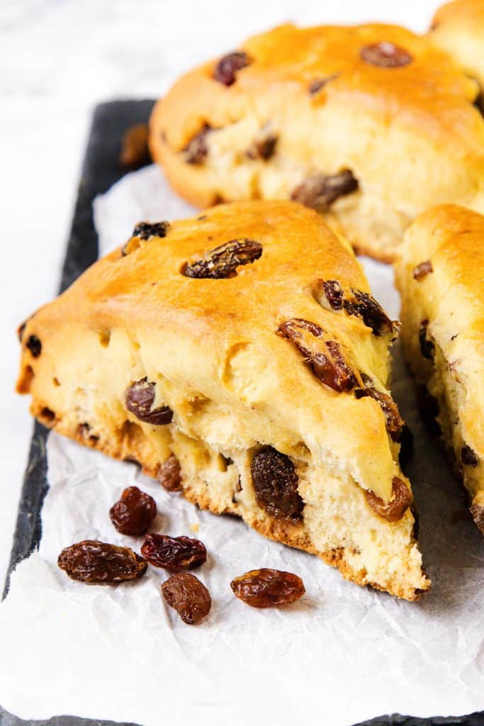 A close-up of a slice of golden-brown raisin scone, studded with plump raisins, rests on crumpled parchment paper. More raisin scones and scattered raisins are visible in the background.