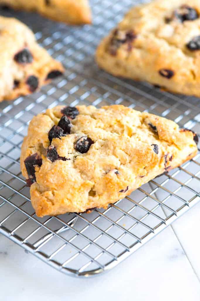 A golden-brown scone with raisins sits on a cooling rack, alongside a few raisin scones. More freshly baked treats are visible in the background on the same rack. The shiny surface indicates their just-out-of-the-oven texture, resting on a light countertop.