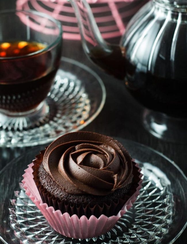 Devil's Food Cupcakes with Dark Chocolate Frosting on a crystal plate and a cup of coffee in the background.