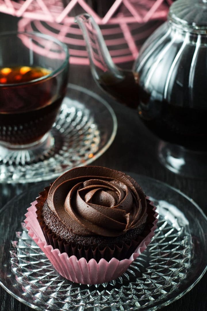 Devil's Food Cupcakes with Dark Chocolate Frosting on a crystal plate and a cup of coffee in the background.