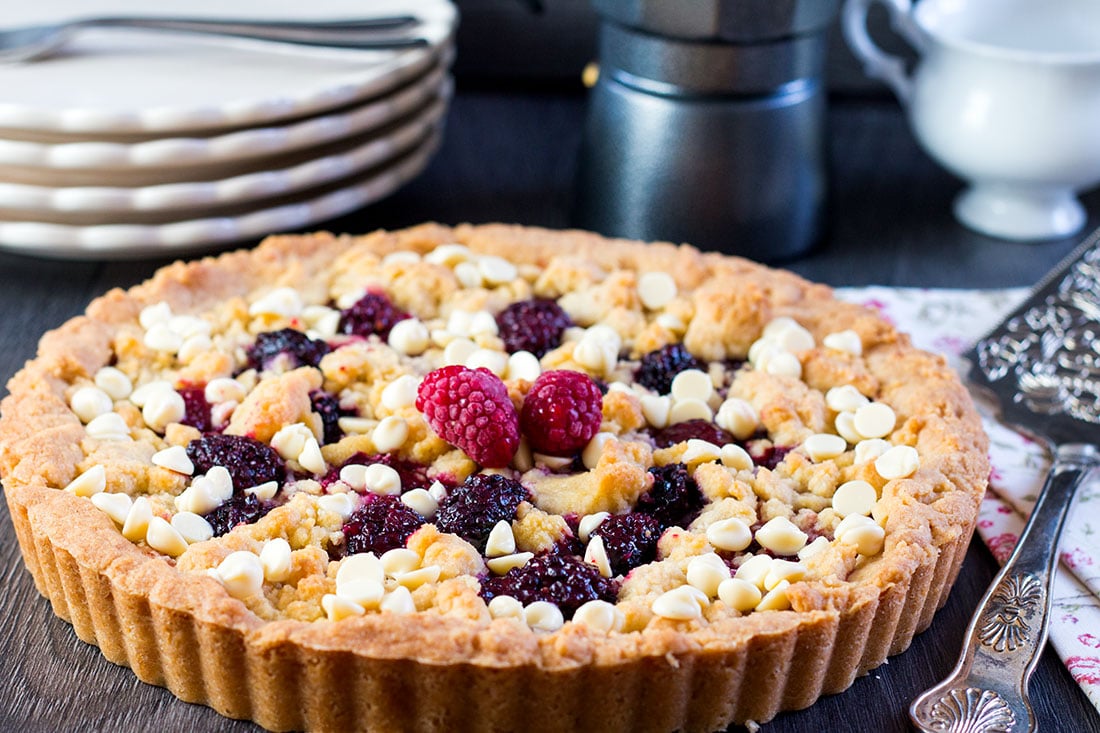 Raspberry and White Chocolate Shortbread Tart decorated with fresh raspberries with cake slice and a stack of plates in the backgroun