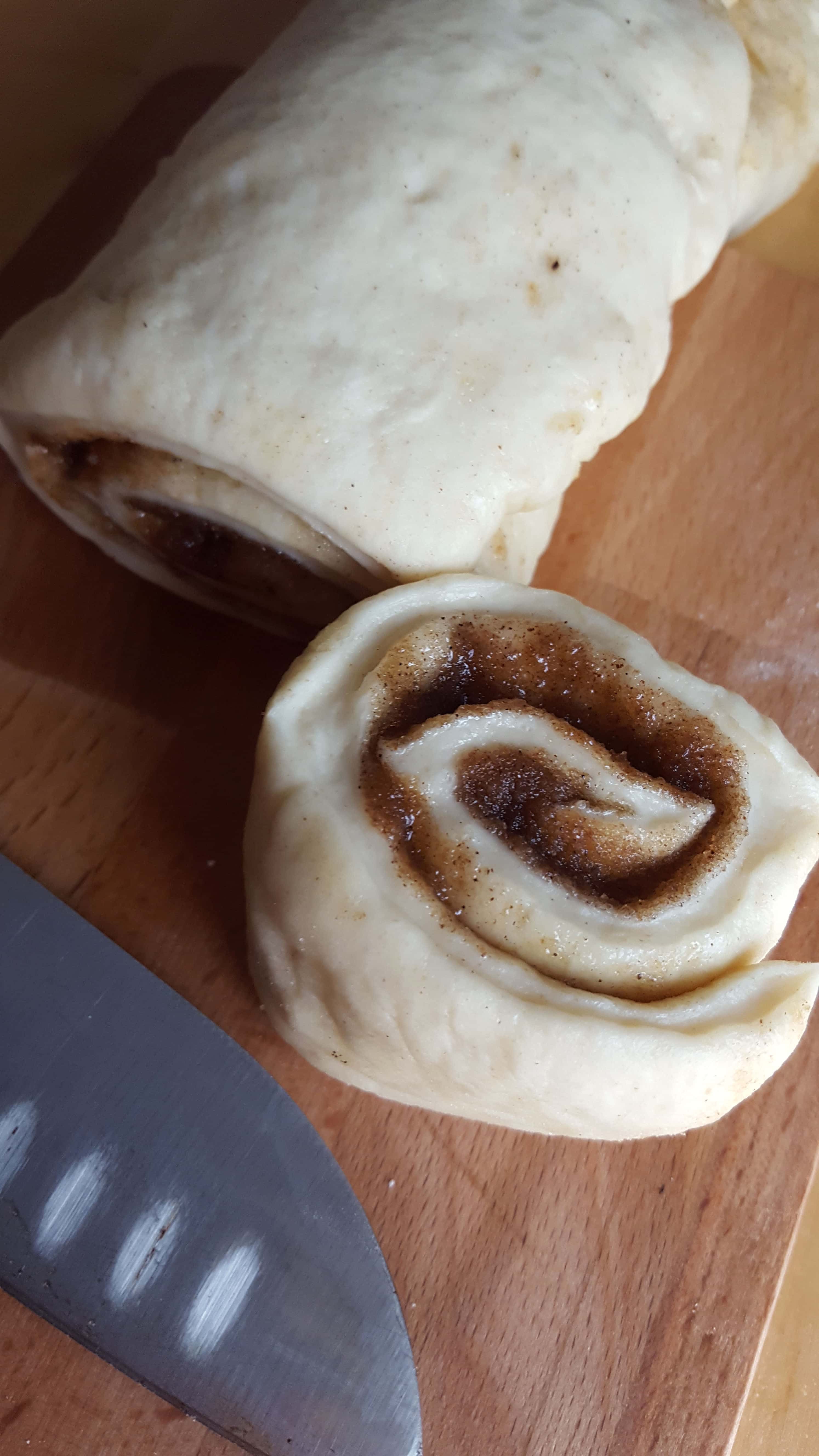 The rolled up dough on a cutting board with one bun cut showing the swirls of cinnamon and a knife next to it.