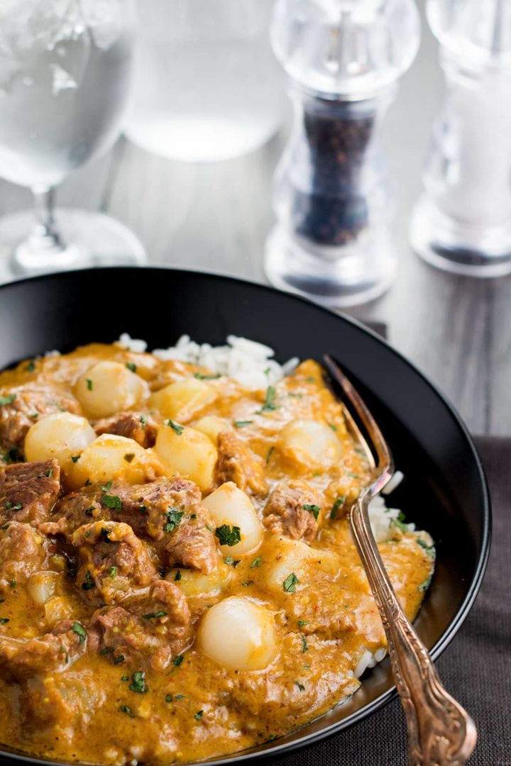 Beef Massaman Thai Curry with rice in a black bowl with a fork, glass of water and salt and pepper mills in the background