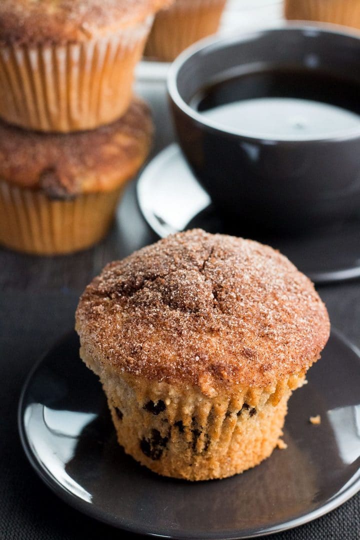 A Cinnamon Raisin Jumbo Muffin Topped with Cinnamon Sugar on a plate with a cup of coffee behind it.