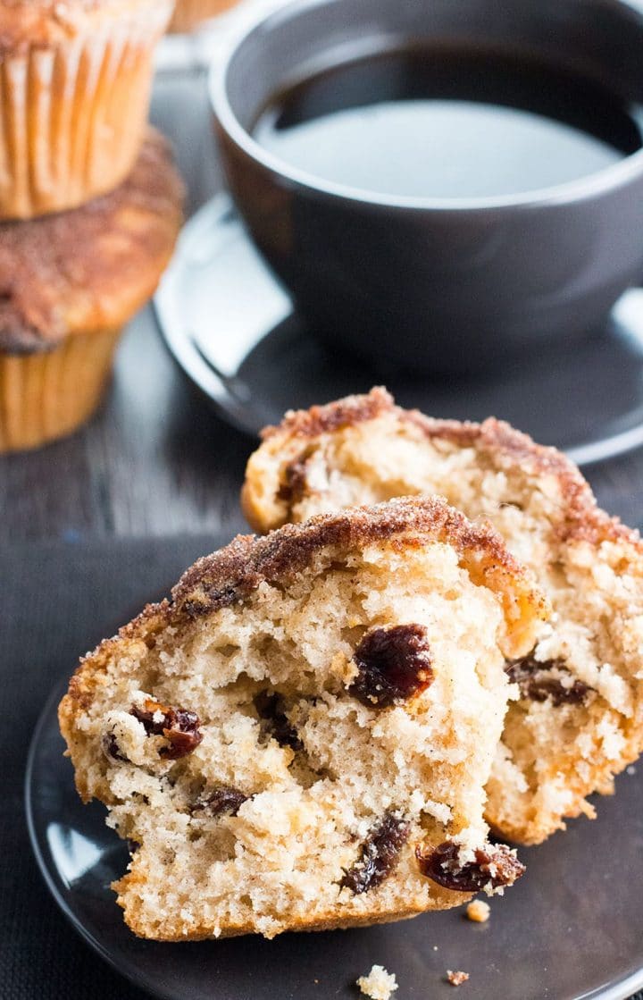 a Cinnamon Raisin Jumbo Muffin Topped with Cinnamon Sugar broken in half on a plate with a cup of coffee in the background