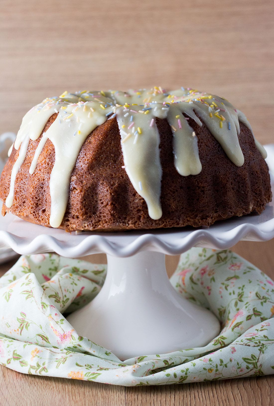 a banana bundt cake on a cake dish drizzled with icing and topped with sprinkles