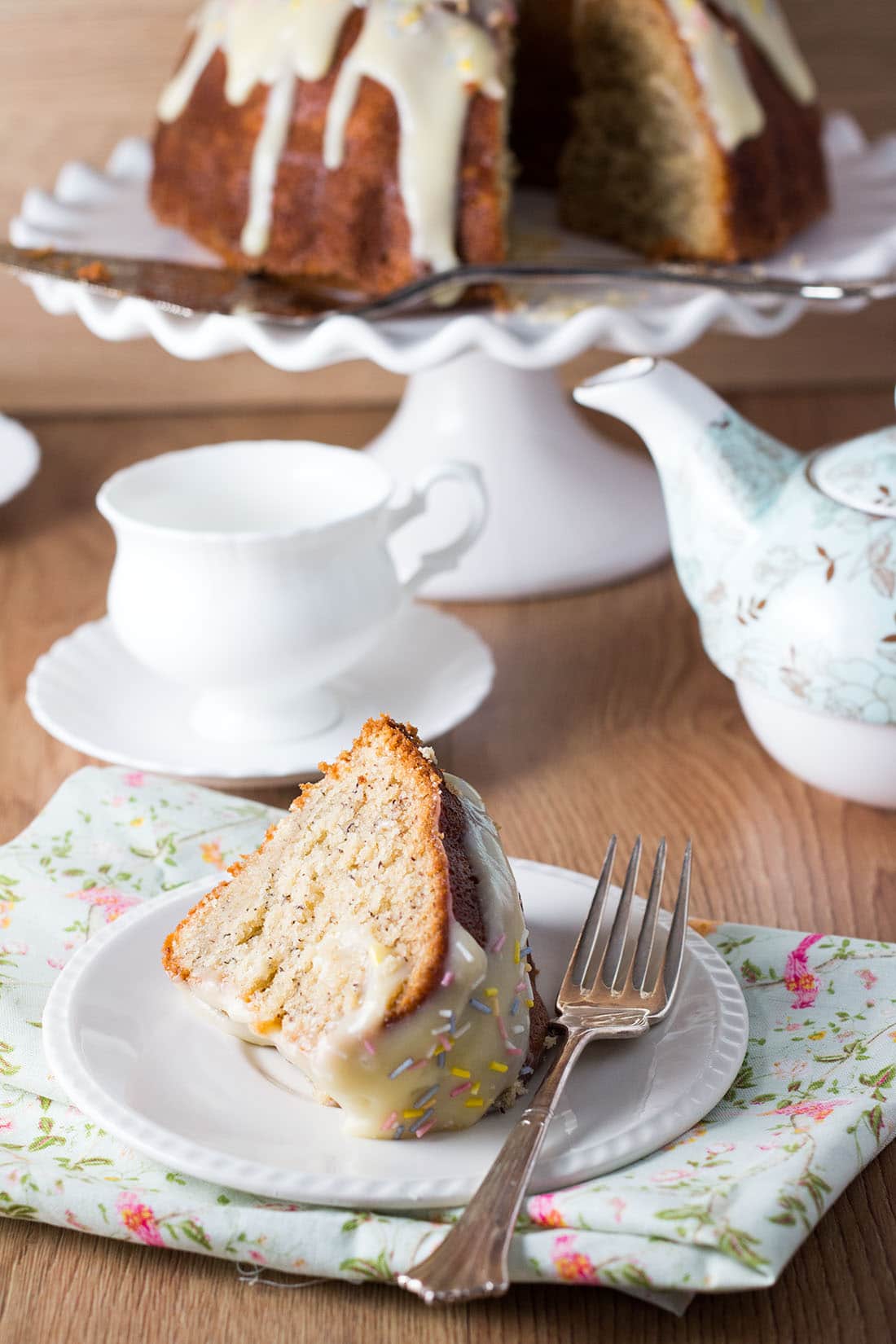 Vanilla Bean Banana Bundt Cake with Cream Cheese Icing sliced on a plate with a fork with a tea pot and cup and saucer in the background