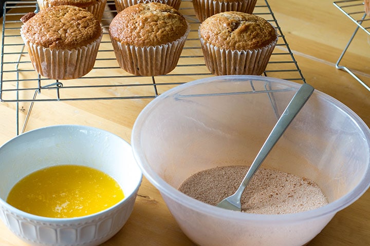the bowls of melted mutter and cinnamon sugar topping on a table with next to the cooling muffins