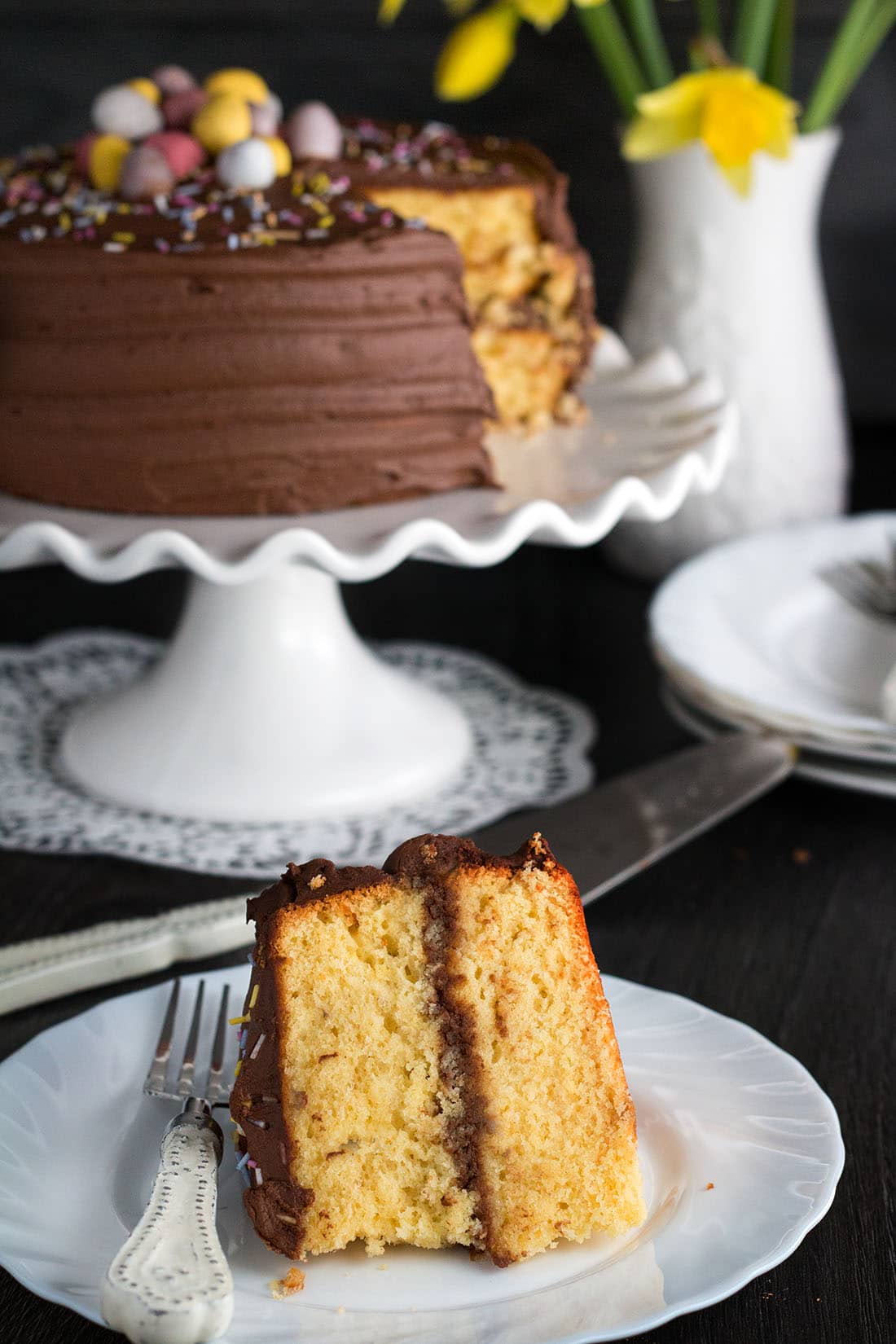 A piece of cake on a plate, with the rest of the cake in the background