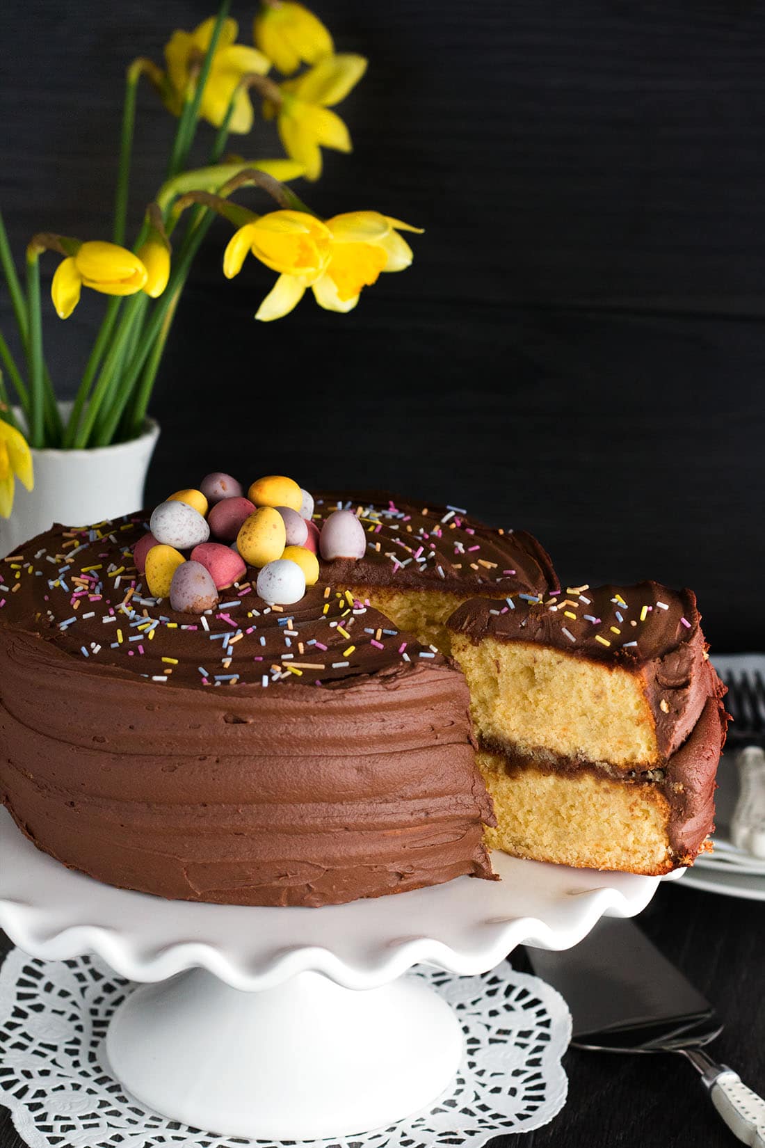 A piece of yellow celebration cake with chocolate frosting being removed from the whole cake, with spring flowers in the background