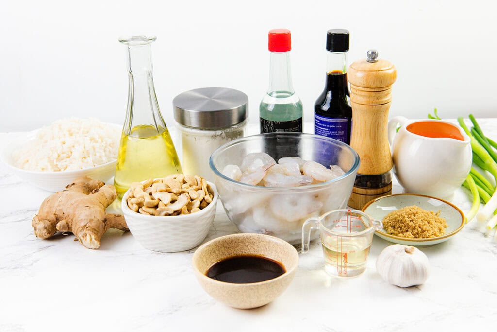 A variety of ingredients on a white countertop, ready for 10 Minute Cashew Shrimp, including a bowl of raw shrimp, cooked rice, cashews, ginger, garlic, green onions, oil, soy sauce, brown sugar, pepper grinder, and sauces in bottles and pitchers.