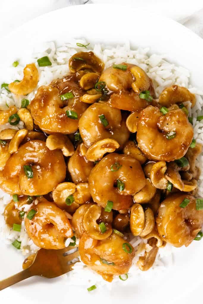 A plate of 10 Minute Cashew Shrimp features white rice topped with shrimp, cashews, and a glossy brown sauce, garnished with chopped green onions. A gold fork rests on the side of the plate.