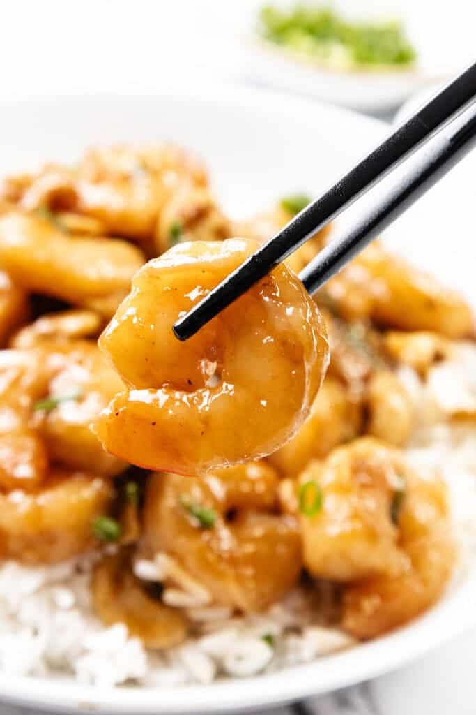 Close-up of black chopsticks holding a saucy shrimp above a plate of 10 Minute Cashew Shrimp served over white rice, garnished with green herbs. The background is softly blurred.