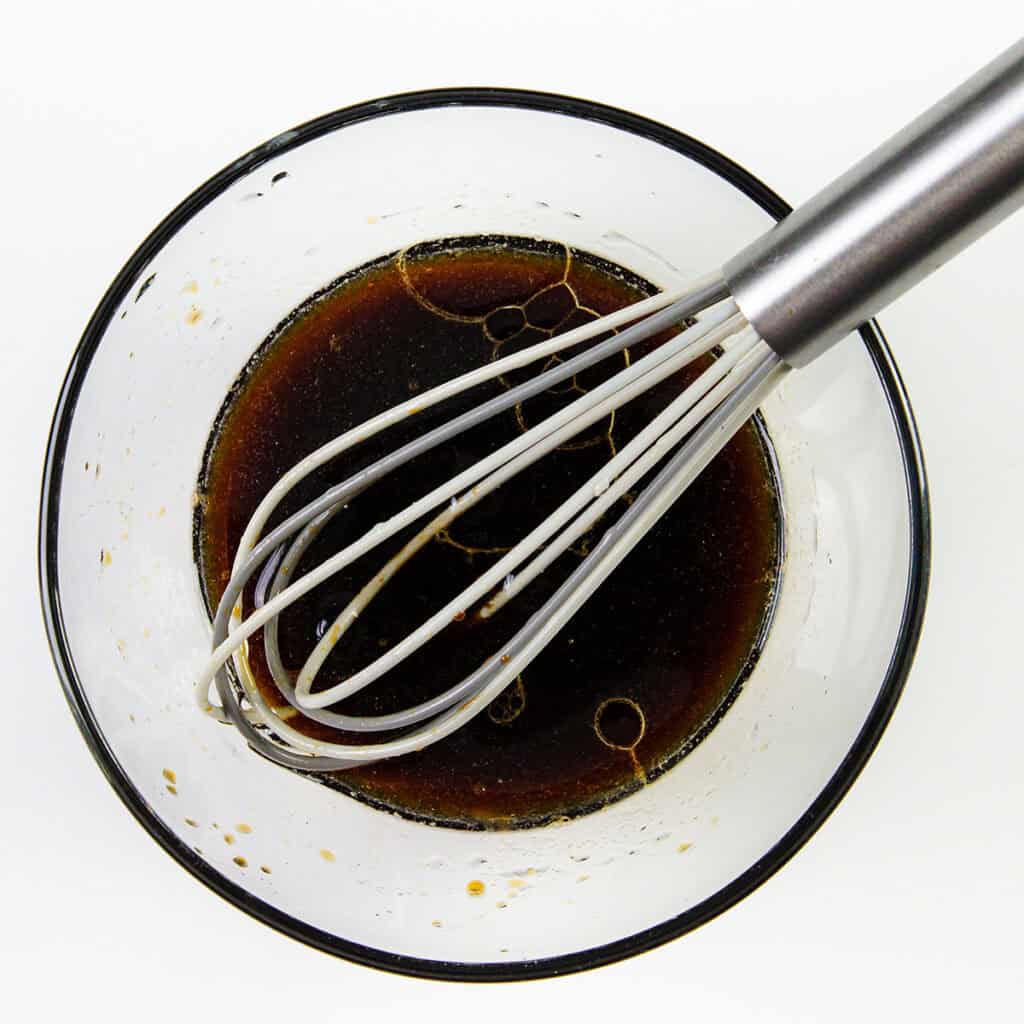 A metal whisk rests in a glass bowl containing a dark brown liquid with small bubbles and oil droplets—perhaps a marinade for 10 Minute Cashew Shrimp—set against a white background.