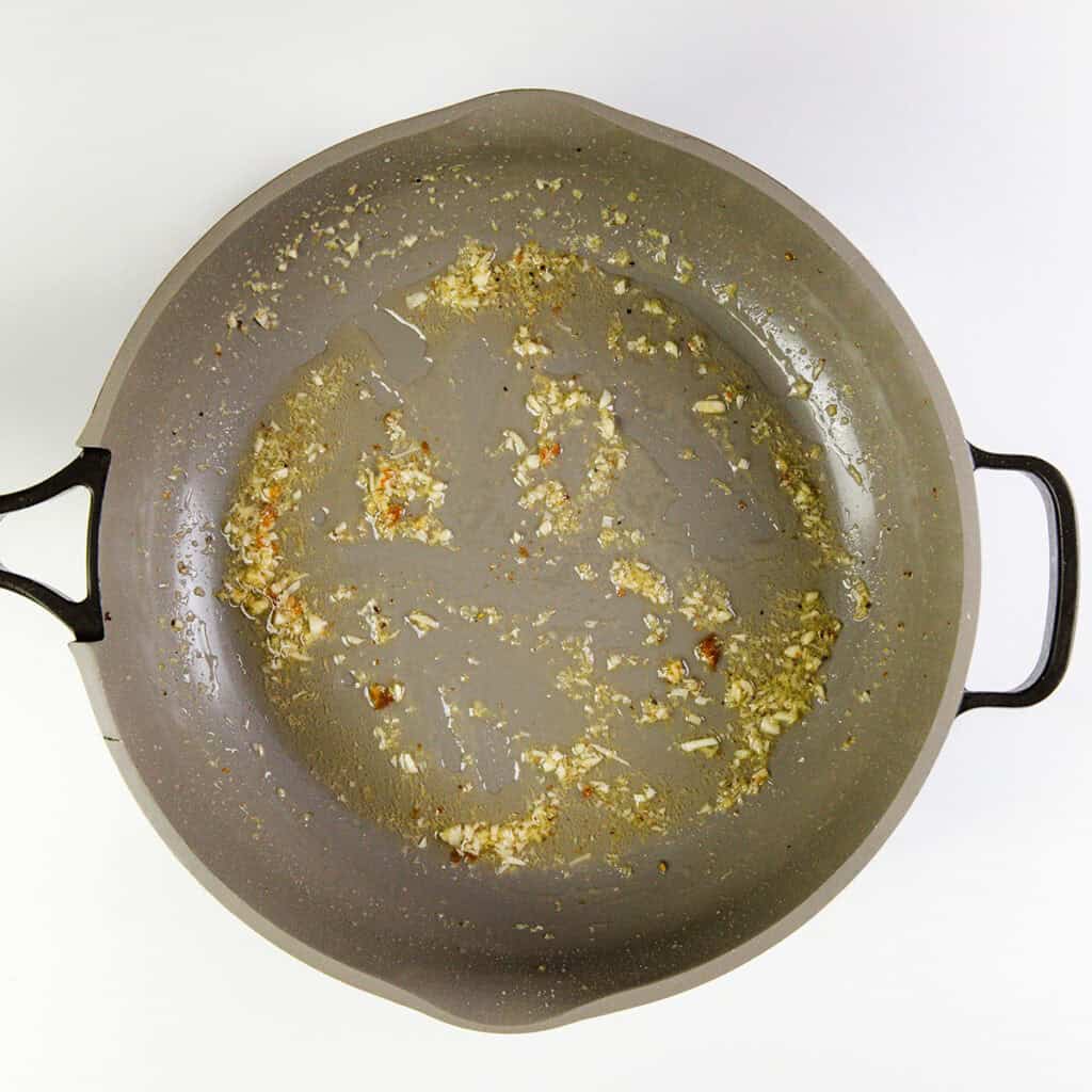 A gray pan with black handles contains bits of sautéed minced garlic and oil spread across its surface, the first step in making 10 Minute Cashew Shrimp. The background is white.