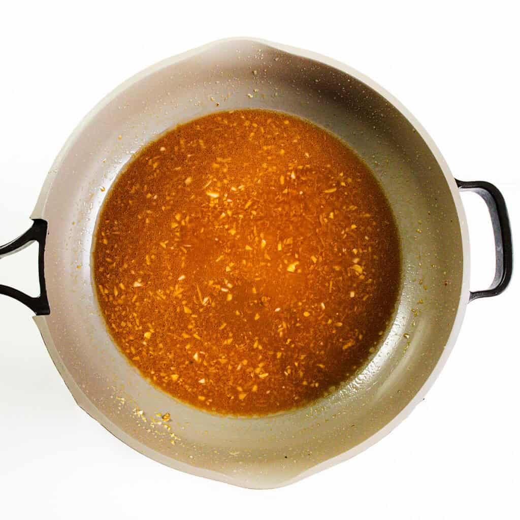 A round gray skillet with black handles contains a simmering brown sauce with visible minced garlic or onion pieces—perfect for making 10 Minute Cashew Shrimp—on a white background.