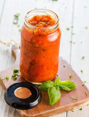 A jar of Easy Homemade Tomato Sauce with basil and chopped parsley scattered around it