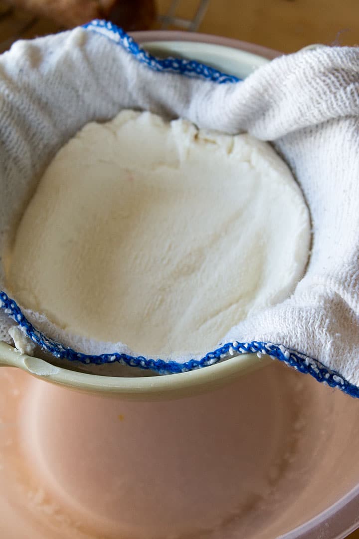 A close up of ricotta cheese in a strainer lined with a cloth and liquid in the bowl beneath