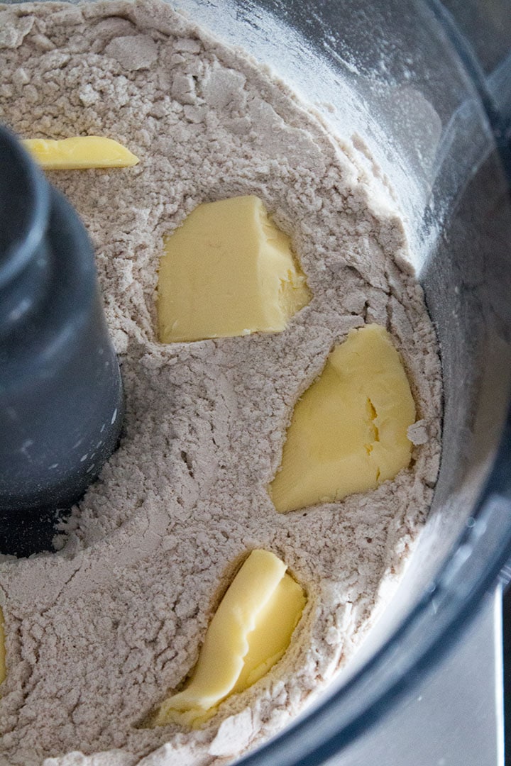 a close up of a food processor bowl with cubed butter laying in the dry ingredients.