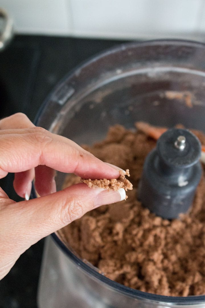 a close up of a food processor bowl with the butter cut into the dry ingredients. with fingers pinching a clump of it together.