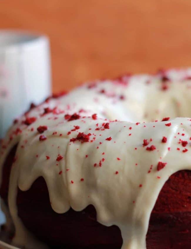 Red Velvet Bundt Cake with Cream Cheese Icing on a cake plate with red crumbs cattered on top