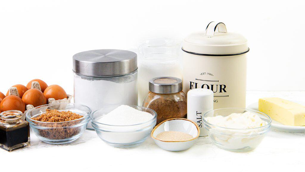 A variety of baking ingredients for Cinnamon Crumb Buns, including eggs, brown and white sugar, flour, salt, butter, yeast, sour cream, vanilla, and baking powder, are arranged in bowls and containers on a white surface.