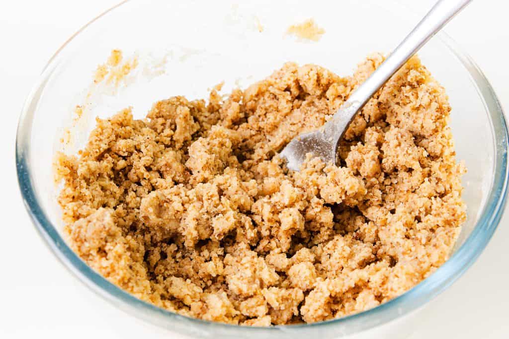 A clear glass bowl filled with crumbly brown sugar and butter mixture, perfect for Cinnamon Crumb Buns, with a metal fork partially buried in the mixture, on a white background.