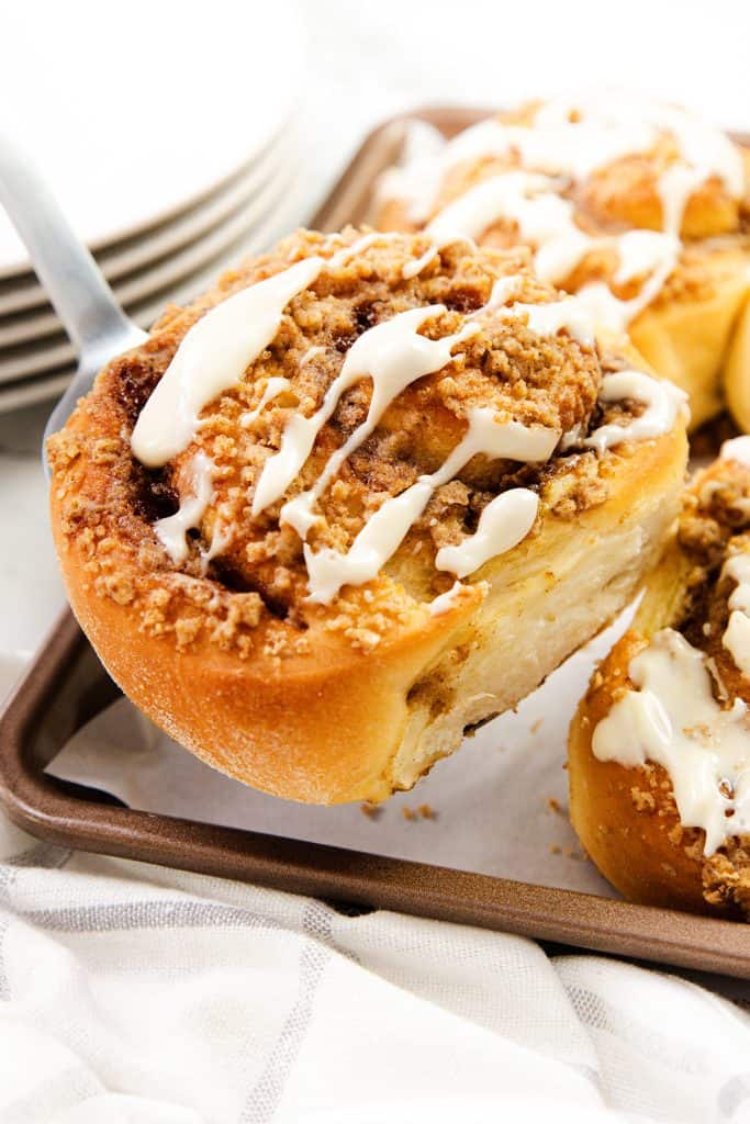 A close-up of Cinnamon Crumb Buns topped with crumbly streusel and drizzled with white icing, being lifted from a baking tray. Other cinnamon rolls and stacked plates are visible in the background.