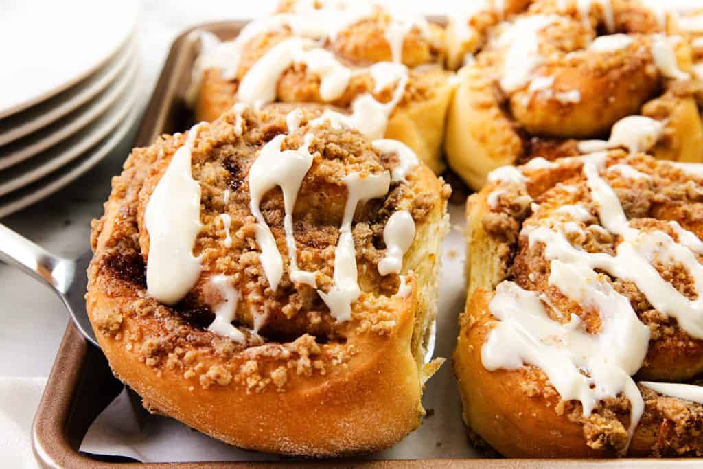 Close-up of Cinnamon Crumb Buns with crumbly streusel topping and drizzled white icing, sitting on a baking tray. Plates are stacked in the background.