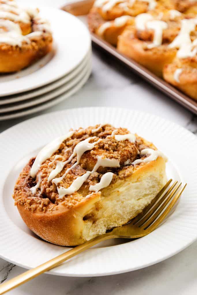 A freshly baked Cinnamon Crumb Bun with crumb topping and white icing drizzle sits on a white plate with a gold fork, while a tray of more Cinnamon Crumb Buns and stacked plates can be seen in the background.