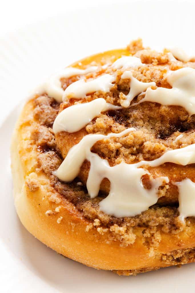A close-up of Cinnamon Crumb Buns topped with crumbly streusel and drizzled with white icing, served on a white plate.