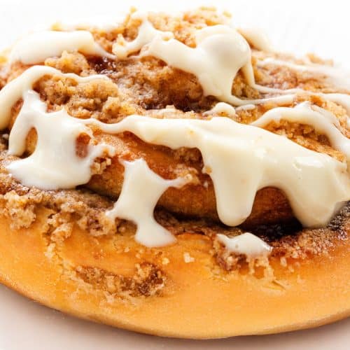 A close-up of Cinnamon Crumb Buns topped with creamy white icing and crumbly brown sugar, served on a white plate.