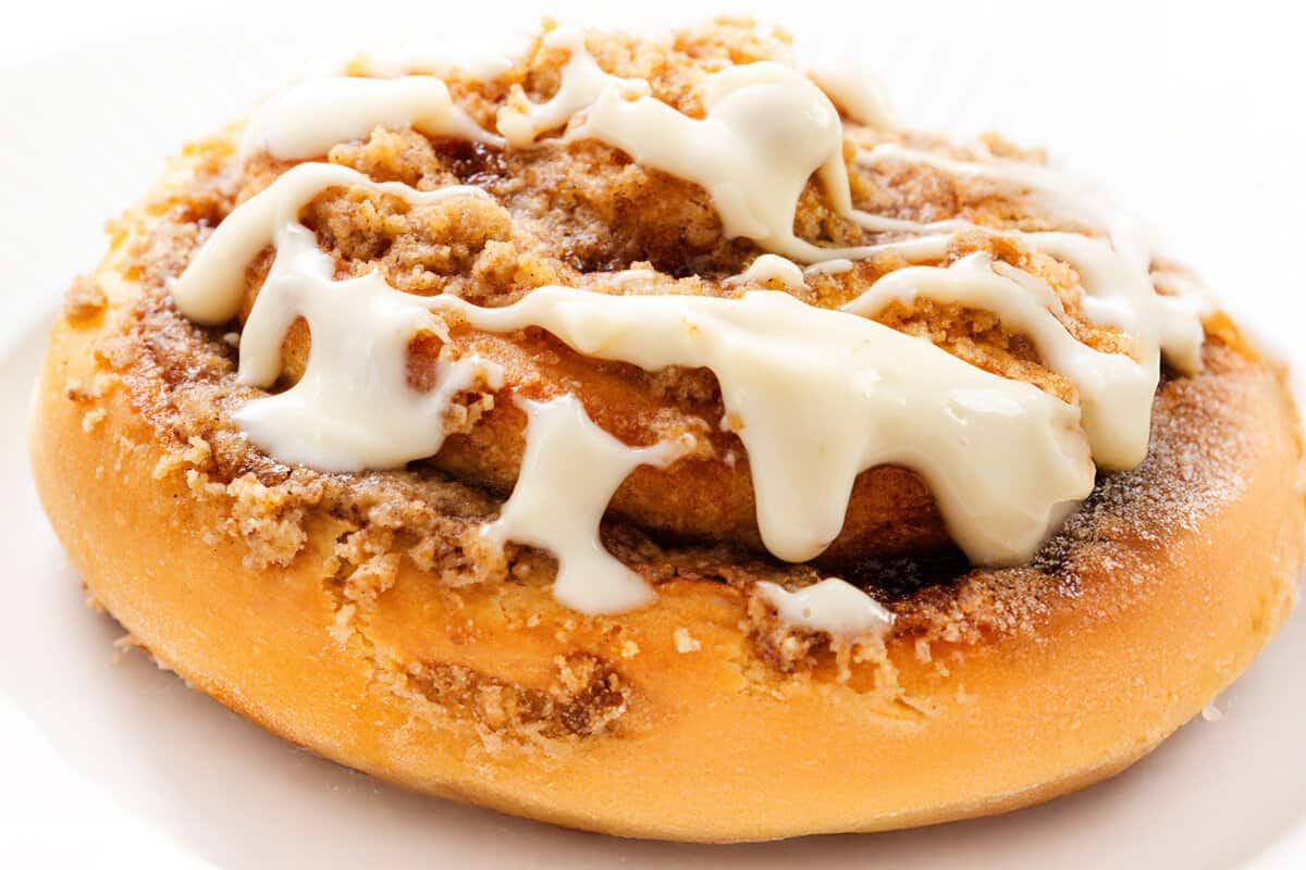 A close-up of Cinnamon Crumb Buns topped with creamy white icing and crumbly brown sugar, served on a white plate.