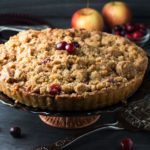 An apple cranberry crumb pie on a pie stand with cranberries on top, scattered on the table and some apples in the background.