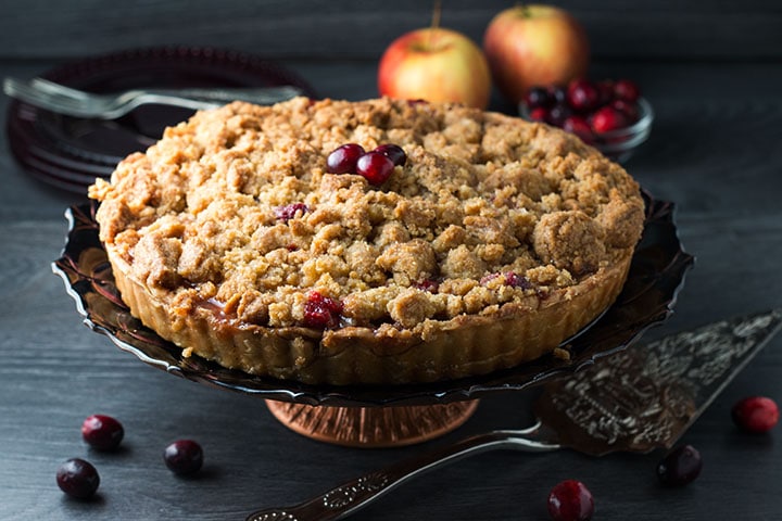 An apple cranberry crumb pie on a pie stand with cranberries on top, scattered on the table and some apples in the background.