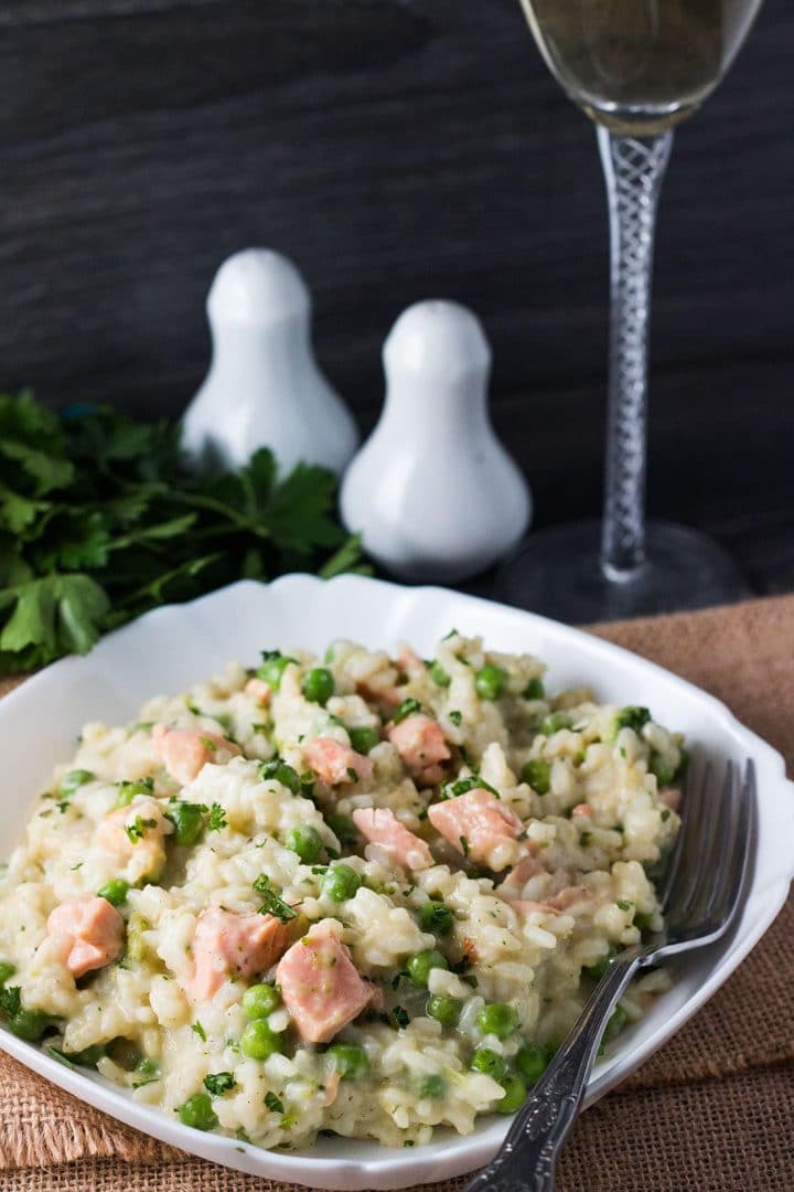 A bowl of salmon pea risotto with a fork and stem of a wine glass, salt and pepper shakers, and fresh herbs in the background