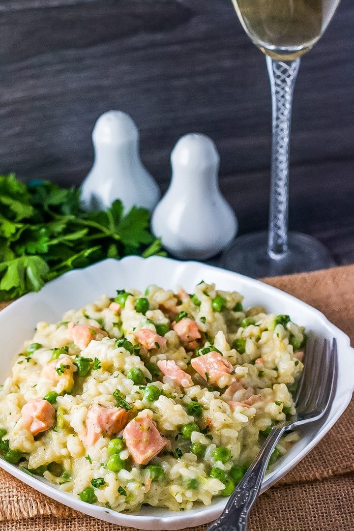 A bowl of salmon pea risotto with a fork and stem of a wine glass, salt and pepper shakers, and fresh herbs in the background