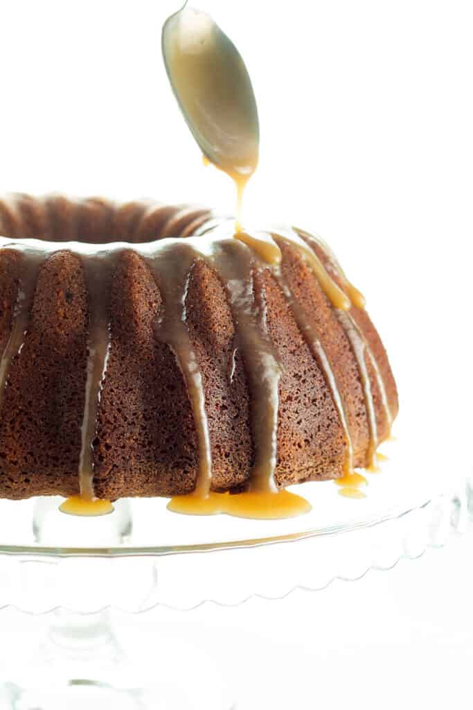 A Brown Sugar Bundt Cake on a glass cake stand being drizzled with Caramel Glaze