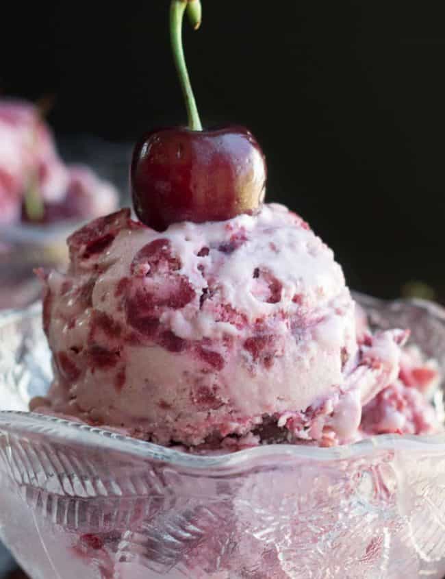 Cherry ice cream in a pretty glass bowl topped with a fresh cherry with more icecream and cherries in the background.